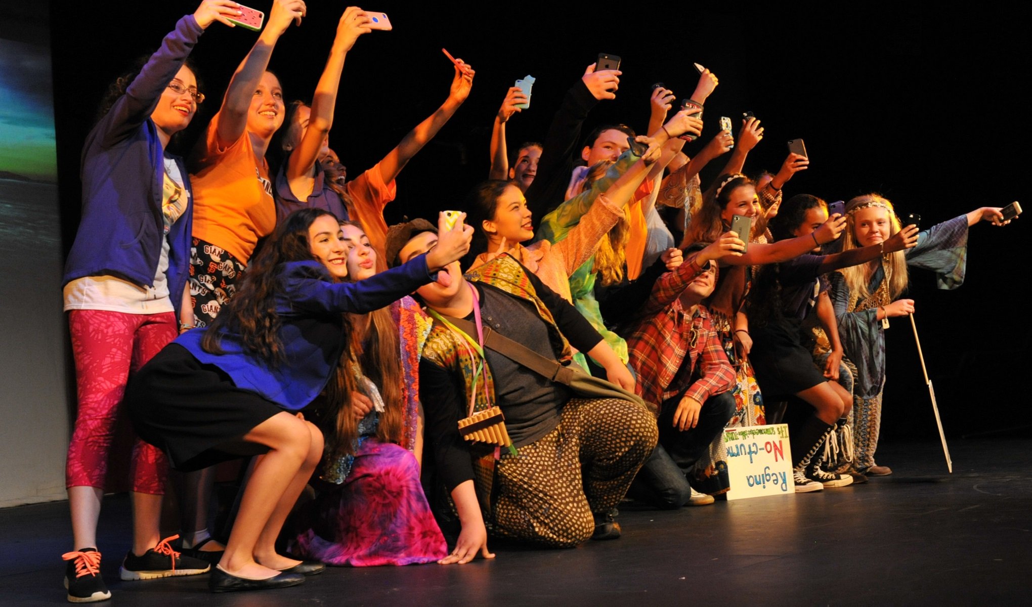 A group of students on stage all taking selfies of themselves.