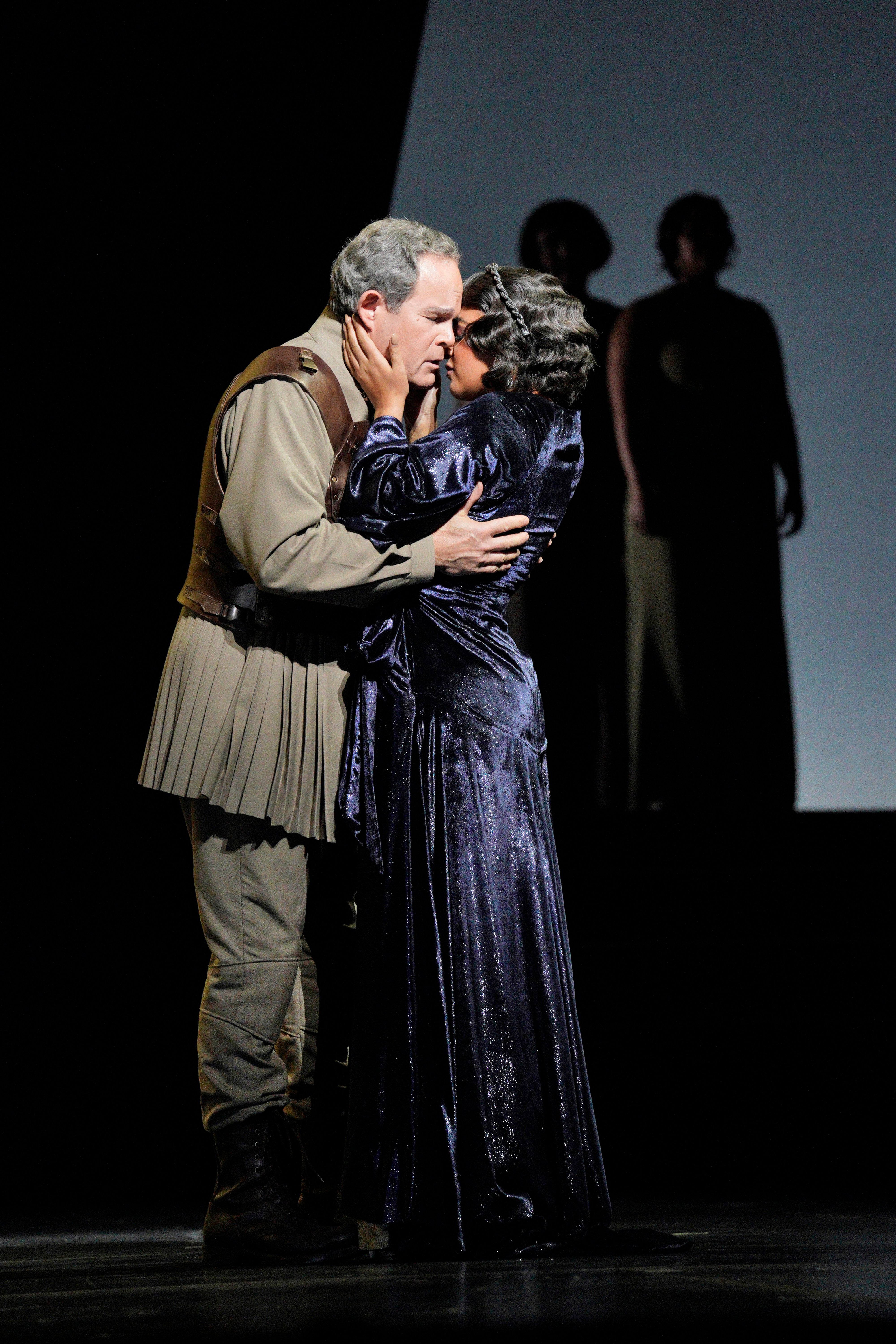 Edris and Finley on stage embrace in an emotional scene, with a dramatic backdrop and two shadowy figures in the background.